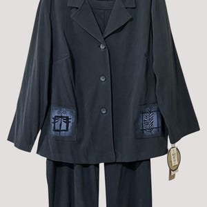 May include: A dark charcoal gray two-piece set consisting of a long-sleeve button-down jacket with a notched collar and matching trousers. The jacket features two front patch pockets, each decorated with a blue square graphic print.