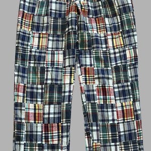 May include: Full-length trousers featuring a patchwork design composed of various plaid fabric squares in shades of navy blue, forest green, red, yellow, and white. The pants include a waistband with a single button closure and belt loops.