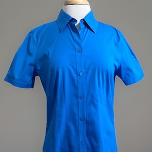 May include: A bright blue short-sleeved button-down shirt with a classic collar and a front placket featuring matching blue buttons. The shirt is displayed on a mannequin, showcasing a tailored fit with vertical seams running down the front.