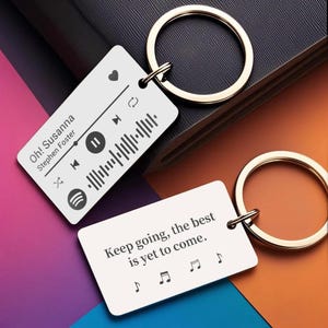 Puede incluir: Dos llaveros rectangulares de metal plateado con anillas. Uno presenta una interfaz de reproductor de música con el texto Oh! Susanna de Stephen Foster y una onda sonora. El otro muestra el texto Keep going, the best is yet to come.