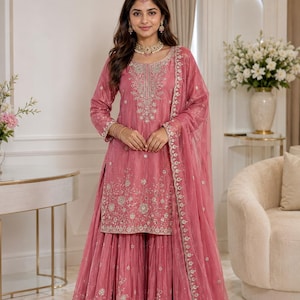 May include: A dusty rose sharara suit featuring a knee-length tunic with silver floral embroidery, a matching flared bottom, and a sheer dupatta with an embroidered border. The ensemble is accented with a gold-toned necklace and headpiece.