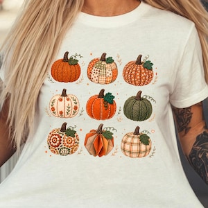 Könnte beinhalten: Ein elfenbeinfarbenes Kurzarm-T-Shirt mit einem Raster aus neun verschiedenen Kürbissen. Die Kürbisse zeigen verschiedene Muster, darunter orange, grün, kariert, floral und strukturiert, jeweils mit einem kleinen grünen Stiel.