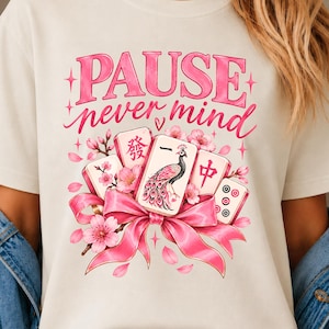 Könnte beinhalten: Ein elfenbeinfarbenes T-Shirt mit einem Grafikdruck und dem Text PAUSE never mind in Pink. Das Design zeigt Mahjong-Steine, eine zentrale Pfauenillustration, rosa Kirschblüten und eine große rosa Schleife am Fuß der Spielsteine.