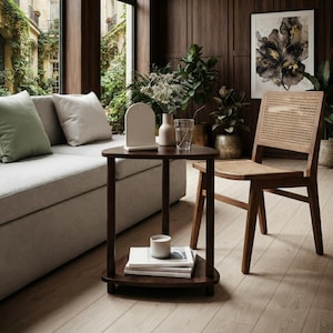 May include: A dark brown wooden side table with a triangular top and a lower shelf. The table features three cylindrical legs and holds a white arched decorative object, a clear glass vase with dried flowers, and a stack of white books.