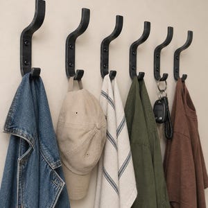 Pode incluir: Um conjunto de seis ganchos de parede de metal preto fosco montados em uma fileira horizontal. Cada gancho apresenta um design curvo de ponta dupla fixado com dois parafusos visíveis. Vários itens, incluindo uma jaqueta jeans e um boné, estão pendurados.