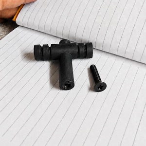 Puede incluir: Un pomo de mueble en forma de T de color negro mate con un mango cilíndrico texturizado y ranurado, junto con un tornillo de cabeza plana a juego con una ranura en forma de estrella. El pomo tiene un orificio roscado central.