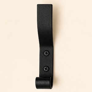 May include: A matte black metal wall hook with a curved upper arm and a rounded lower hook. The vertical mounting plate features two visible black Phillips head screws aligned vertically in the center of the lower section of the hook.