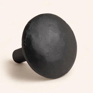 May include: A matte black cabinet knob with a circular, slightly domed head and a textured, hammered surface. The knob features a cylindrical base extending from the center of the back, designed for attachment to furniture or cabinetry doors.