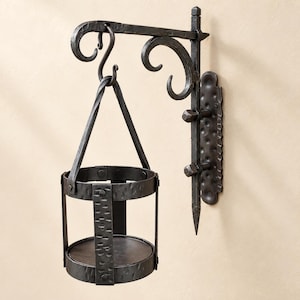 Puede incluir: Un soporte de pared de metal martillado gris oscuro con un diseño de volutas decorativas y una placa de montaje vertical. Un soporte cilíndrico de metal a juego con acabado texturizado cuelga del brazo mediante un gancho central y correas.