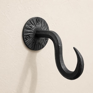 Puede incluir: Un gancho de pared de metal forjado a mano en color gris oscuro, con una placa de montaje circular que presenta un patrón texturizado similar a rayos de sol. El gancho tiene un diseño curvo y cónico, fijado con dos tornillos negros visibles.