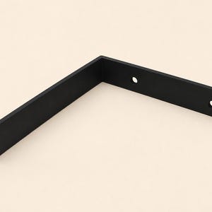 Può includere: Una staffa per mensola a forma di L in metallo nero opaco con un design piatto e angolare, dotata di due fori di montaggio circolari preforati posizionati lungo uno dei bracci. La staffa presenta una finitura liscia e un angolo retto.