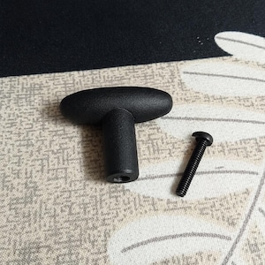 May include: A matte black T-shaped cabinet knob with a textured finish and a single matching black metal mounting screw. The knob features a horizontal handle and a cylindrical base with a threaded hole at the bottom for installation purposes.