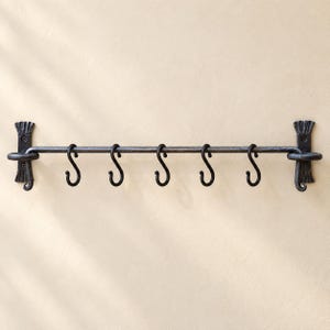 May include: A dark gray metal wall-mounted rack featuring a horizontal rod supported by two decorative brackets at each end. Five removable S-shaped hooks are suspended from the rod, providing multiple points for hanging various items.