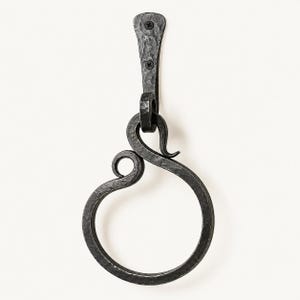 Könnte beinhalten: Ein dunkelgrauer, handgeschmiedeter Handtuchring aus Eisen mit einer texturierten, gehämmerten Oberfläche. Der kreisförmige Ring hängt an einer dekorativen, geschwungenen Metallhalterung mit zwei sichtbaren schwarzen Schrauben.
