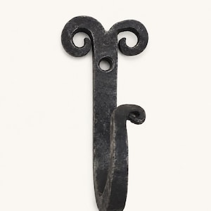 May include: A dark gray, hand-forged metal wall hook featuring a textured, matte finish. The top section displays two symmetrical, outward-curling scrolls with a central circular mounting hole, while the lower portion forms a single upward-curving hook.