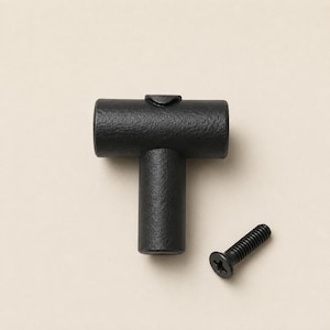 Puede incluir: Un pomo para mueble en forma de T de color negro mate con un acabado de superficie texturizado. El pomo presenta una barra cilíndrica horizontal unida a una base cilíndrica vertical. Se incluye un tornillo de metal negro con cabeza plana.