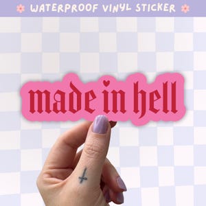 Op de afbeelding: Een waterbestendige vinylsticker met de tekst made in hell in een donkerroze gotisch lettertype op een lichtroze achtergrond. De sticker heeft een gestanste vorm die de contouren van de letters volgt, inclusief een klein hartje boven de letter i.