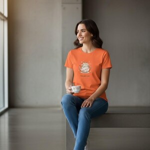 May include: An orange short-sleeved t-shirt featuring a graphic of a cat sitting inside a floral teacup with the text 'tea pot cat' printed below the illustration, paired with blue denim jeans and white trainers.