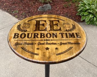 Pode incluir: Uma mesa redonda de madeira com acabamento brilhante, apresentando as letras EE, o texto BOURBON TIME e a frase Good Friends - Good Bourbon - Great Memories. O tampo tem dois ícones de barril, três estrelas e uma base de pedestal preta.