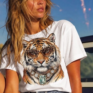 Könnte beinhalten: Ein weißes Kurzarm-T-Shirt mit einem Grafikdruck eines Tigers, der eine Brille mit Leopardenmuster und eine Halskette mit einem großen smaragdgeschliffenen Edelstein trägt, kombiniert mit hellblauen Jeansshorts und cremefarbenen Leder-Cowboystiefeln.