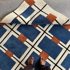 Peut inclure: Un tapis touffeté présentant un motif géométrique composé de rectangles bleus, de petits carrés orange brûlé et de bordures crème, le tout souligné par de fines lignes noires. Un coin du tapis est replié pour révéler l'envers et la texture.