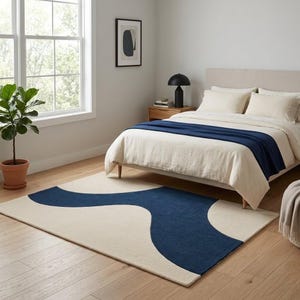 Puede incluir: Una alfombra rectangular con un llamativo patrón de ondas azul oscuro sobre un fondo de color crema. La alfombra está colocada sobre un suelo de madera clara en un dormitorio, complementando la manta azul sobre la cama.