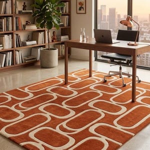 May include: A rectangular area rug featuring a repeating geometric pattern of burnt orange and cream-coloured interlocking rounded rectangles. The rug has a low-pile texture and covers a portion of the floor in a room with a desk and bookshelf.