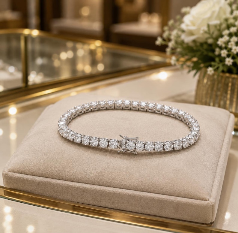 May include: A silver-toned tennis bracelet featuring a continuous row of round, clear, faceted stones set in a metallic frame. The bracelet is secured with a rectangular box clasp mechanism, showcasing a uniform design of sparkling circular links.
