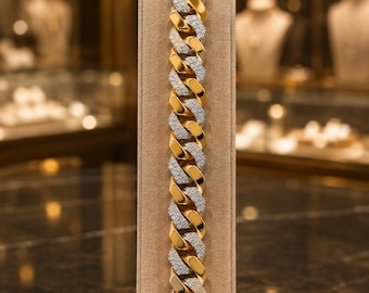 May include: A cuban link chain bracelet featuring alternating polished gold-tone metal links and silver-tone links encrusted with small clear stones. The bracelet is displayed vertically on a beige velvet stand, showcasing a secure box clasp closure.