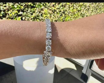 May include: A silver-toned tennis bracelet featuring a continuous row of round, clear, faceted stones set in a metallic frame. The bracelet is secured with a visible box clasp mechanism, showcasing a uniform design of sparkling, circular stones.