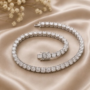 May include: A silver-toned tennis bracelet featuring a continuous row of round, clear, faceted stones set in a metallic frame. The bracelet includes a secure box clasp mechanism with a hinged safety latch, showcasing a polished finish throughout.