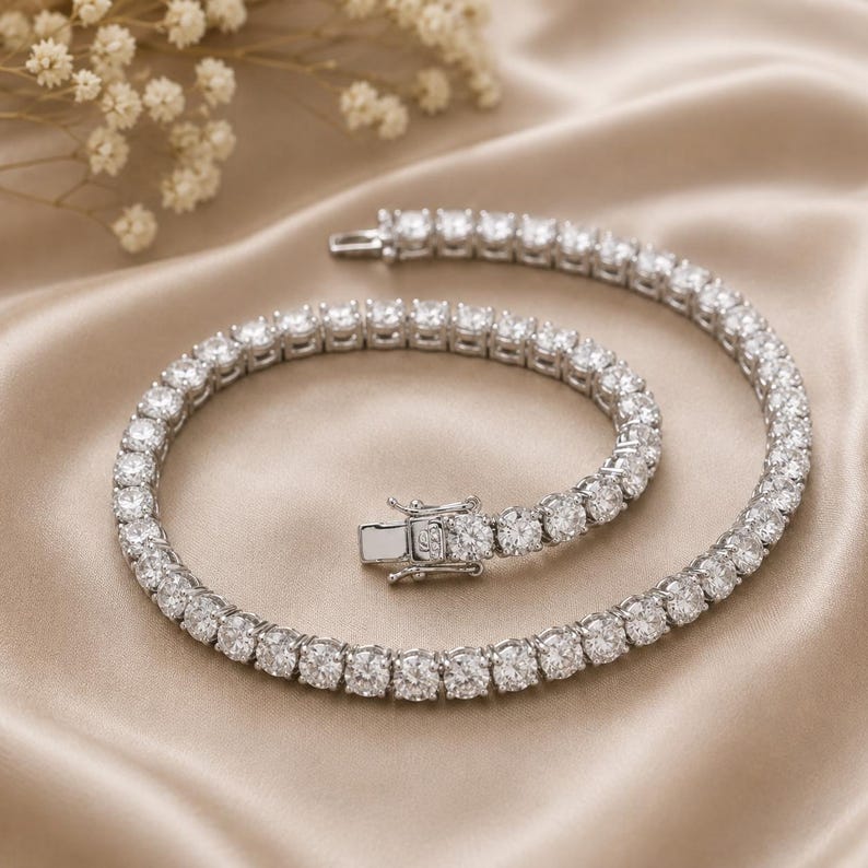 May include: A silver-toned tennis bracelet featuring a continuous row of round, clear, faceted stones set in a metallic frame. The bracelet includes a secure box clasp mechanism with a hinged safety latch, showcasing a polished finish throughout.