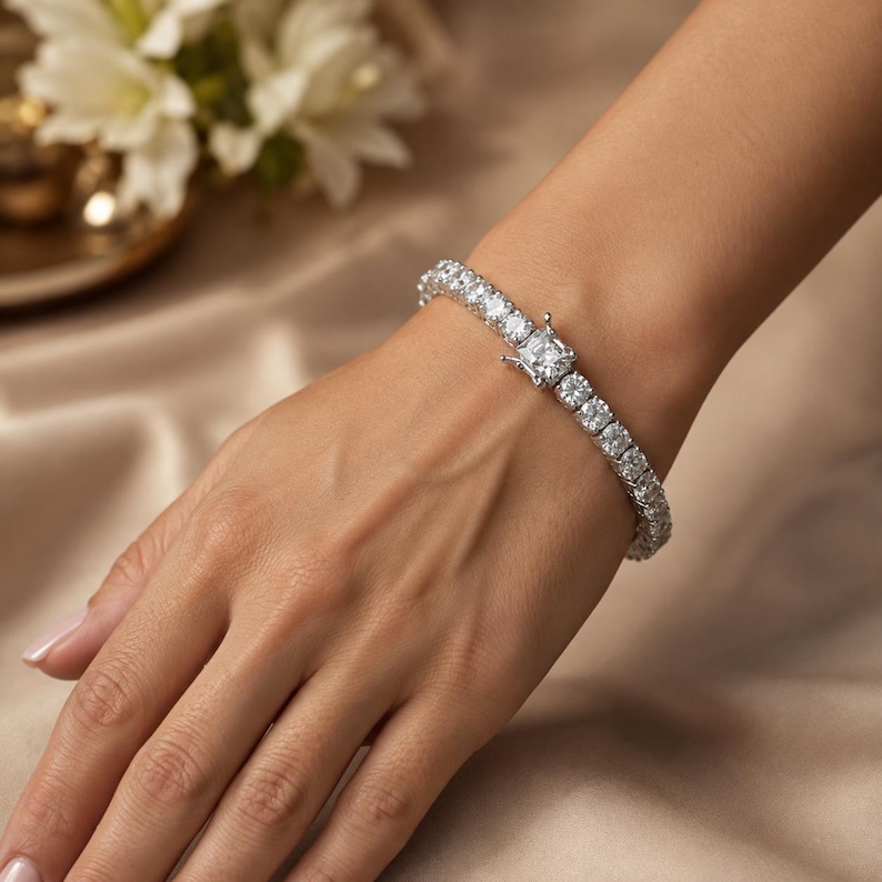 May include: A silver-toned tennis bracelet featuring a continuous row of round, clear, faceted stones. The bracelet includes a prominent square-shaped stone at the clasp mechanism, which secures the piece with a visible metal hinge and latch.