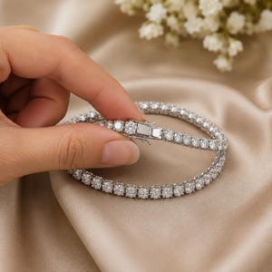 May include: A silver-toned tennis bracelet featuring a continuous row of round, clear, faceted stones set in a metallic frame. The bracelet includes a rectangular box clasp mechanism with a secure side latch, showcasing a polished finish.
