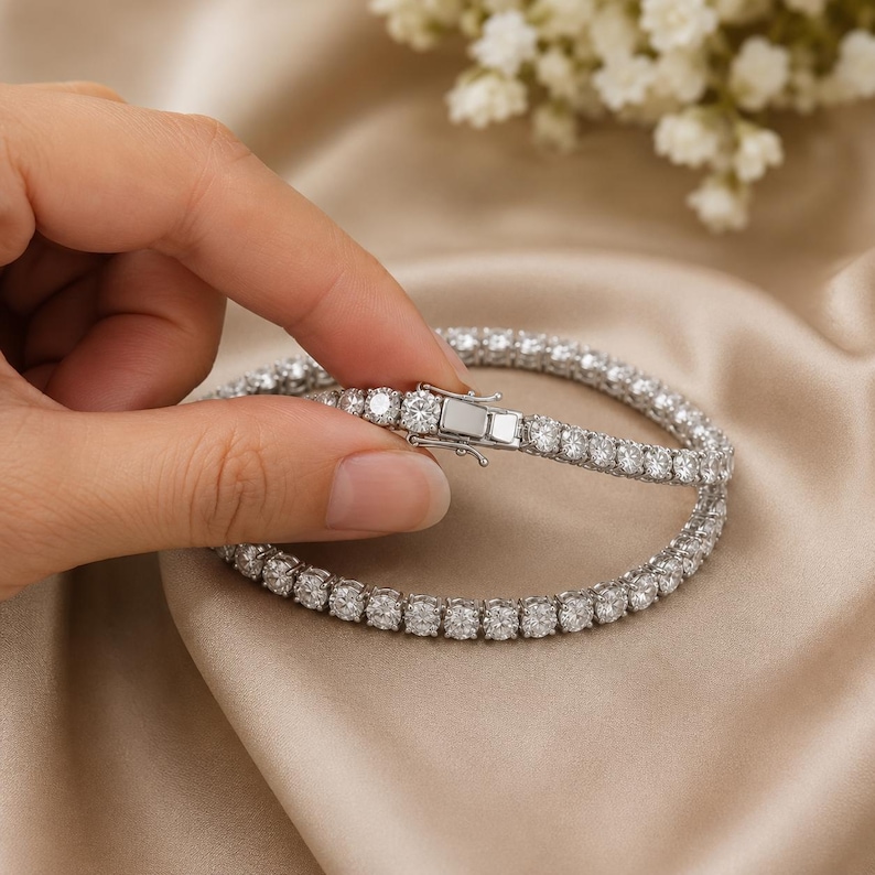 May include: A silver-toned tennis bracelet featuring a continuous row of round, clear, faceted stones set in a metallic frame. The bracelet includes a rectangular box clasp mechanism with a secure side latch, showcasing a polished finish.