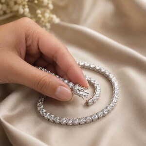 May include: A silver-toned tennis necklace featuring a continuous row of round, clear, faceted stones set in a metallic frame. The necklace includes a secure box clasp mechanism with a side safety latch, showcasing a polished, reflective finish.