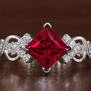 Puede incluir: Un anillo de tono plateado con una gran piedra preciosa de color rojo intenso y corte cuadrado en el centro. La banda está adornada con pequeñas piedras claras de corte redondo y patrones metálicos entrelazados a ambos lados.