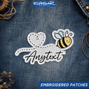 May include: An embroidered patch featuring a yellow and black bee, a white heart outline, and a dashed trail leading to the word Anytext in black cursive script. The patch is displayed against a dark blue denim fabric background.