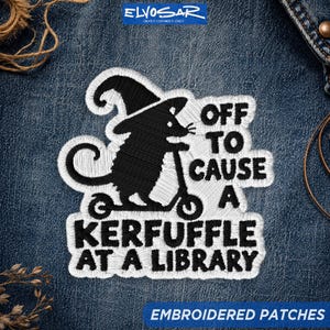 May include: An embroidered patch featuring a black silhouette of a mouse wearing a witch hat and riding a scooter. The text reads OFF TO CAUSE A KERFUFFLE AT A LIBRARY. The design is rendered in black thread with a white border on a blue denim fabric.
