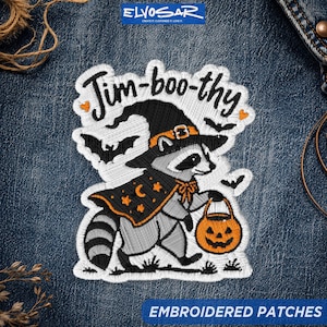 May include: An embroidered patch on blue denim featuring a raccoon wearing a witch hat and a star-patterned cape. The raccoon holds an orange jack-o'-lantern bucket. The text "Jim-boo-thy" is stitched above the raccoon, surrounded by small bats.