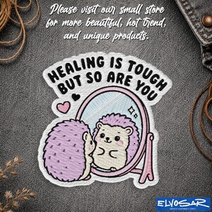 May include: An embroidered patch featuring a light purple hedgehog standing before a pink oval mirror. The hedgehog is reflected in the mirror. The text HEALING IS TOUGH BUT SO ARE YOU is stitched in black above the mirror with a small pink heart.