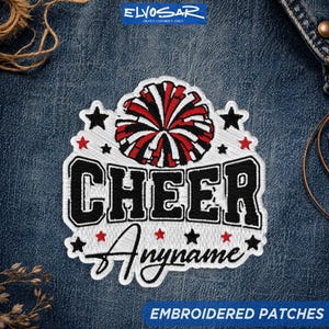 May include: An embroidered patch featuring a red, white, and black pom-pom graphic above the word CHEER in bold black letters. Below, the word Anyname is written in black script. The design includes small black and red stars on a white background.
