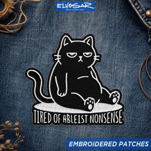 May include: An embroidered patch featuring a black cat with a grumpy expression sitting down. The cat is outlined in white thread. Below the cat, the text reads TIRED OF ABLEIST NONSENSE in white capital letters. The patch is on blue denim fabric.