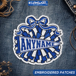May include: An embroidered patch featuring a blue and white cheerleader pom-pom design topped with a blue bow. A triangular pennant is centered across the pom-pom with the text ANYNAME in bold blue capital letters on a white background.