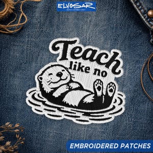 May include: An embroidered patch on blue denim fabric featuring a black and white illustration of an otter floating on its back. The text above the otter reads "Teach like no" in a bold, black, cursive-style font with white stitching details.