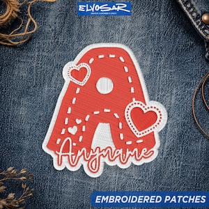 May include: A red embroidered patch in the shape of the letter A, featuring white stitched heart accents and a white border. The patch includes the word Anyname in white cursive script at the bottom, set against a dark blue denim fabric background.