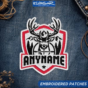 May include: An embroidered patch featuring a black and white deer head with antlers centered within a shield shape outlined in red. The design includes mountain silhouettes and the text ANYNAME in black block letters above a small black star.