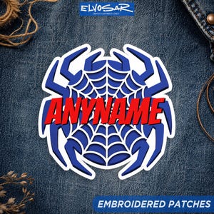 Peut inclure: Un écusson brodé personnalisé présentant un motif de toile d'araignée bleu stylisé avec une bordure blanche. Le mot ANYNAME est imprimé en lettres capitales rouges audacieuses au centre de la toile sur un tissu en jean bleu foncé.