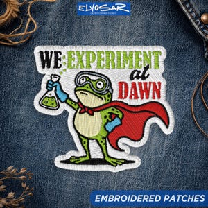 May include: An embroidered patch featuring a green frog wearing safety goggles and a red cape. The frog holds a glass flask with green liquid. The text reads WE EXPERIMENT at DAWN in black, green, and red thread against a white background border.