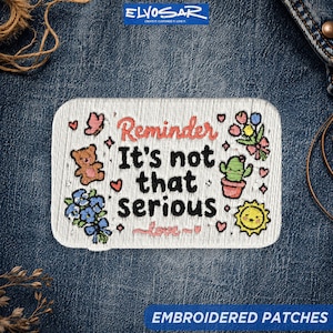 May include: A rectangular embroidered patch with a white background and black text that reads Reminder It's not that serious. The design features a brown teddy bear, a green cactus in a pot, blue flowers, a yellow sun, and small pink hearts.